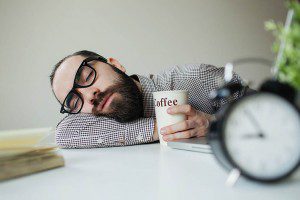 Man sleeps in office on table over laptop with coffee in hand